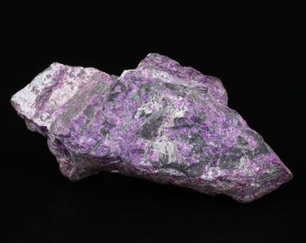 Könnte beinhalten: Ein rohes, unpoliertes Mineralexemplar mit einer leuchtend magentafarbenen und violetten Kristallstruktur, durchsetzt mit einer dunkelgrauen und schwarzen Gesteinsmatrix. Der Stein hat eine unregelmäßige, gezackte Form mit rauer Oberfläche.