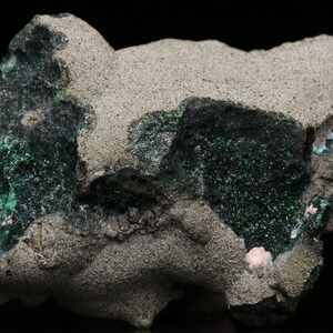 Puede incluir: Una roca gris rugosa con depósitos minerales verdes y rosados. El mineral verde es de un verde brillante y vibrante, mientras que el mineral rosado es de un rosa pálido y suave.