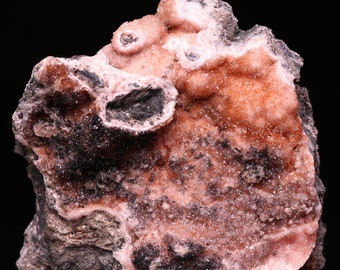 Può includere: Un esemplare minerale rosa e grigio con una superficie ruvida e testurizzata. Il minerale ha una struttura cristallina e sembra essere una combinazione di diversi tipi di minerali.