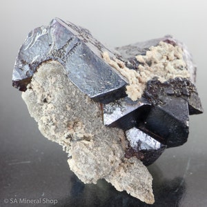 May include: A cluster of sharp, black, metallic crystals with a purplish hue, set against a white, rough, and textured matrix.