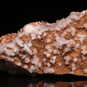 May include: A cluster of white calcite crystals on a brown rock matrix. The crystals are small and have a glassy luster. The rock is rough and textured.