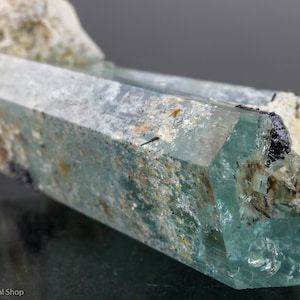 May include: A close-up of a light blue aquamarine crystal with a hexagonal shape. The crystal has a translucent quality with visible internal structures and is partially embedded in a rough, beige matrix. Small black mineral inclusions are present.