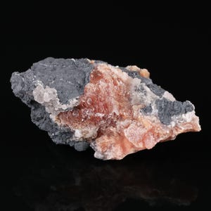 Pode incluir: Um espécime mineral bruto com uma matriz cinzento-escura de textura mate, contrastada por um aglomerado central de formações cristalinas translúcidas cor de pêssego com uma superfície rugosa e granular e pequenos depósitos minerais brilhantes.