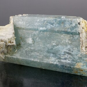 May include: A light blue aquamarine crystal specimen with a rectangular shape. The crystal has a translucent appearance with visible internal structures. The ends of the crystal are rough and have a white, beige, and black mineral composition.