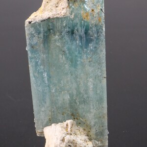 May include: A translucent aquamarine crystal with a rectangular shape. The crystal has a light blue hue with areas of white and black. The base of the crystal has a rough, white, and beige texture. The mineral is displayed against a dark background.