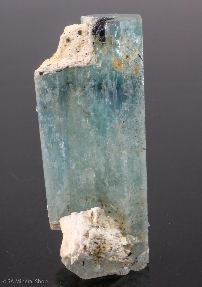May include: A translucent aquamarine crystal with a rectangular shape. The crystal has a light blue hue with areas of white and black. The base of the crystal has a rough, white, and beige texture. The mineral is displayed against a dark background.