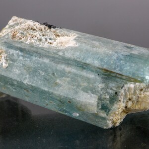 May include: A raw, aquamarine crystal with a rectangular shape. The crystal has a translucent, light blue hue with visible striations. White and beige mineral deposits are present on the edges and ends, with small black inclusions.