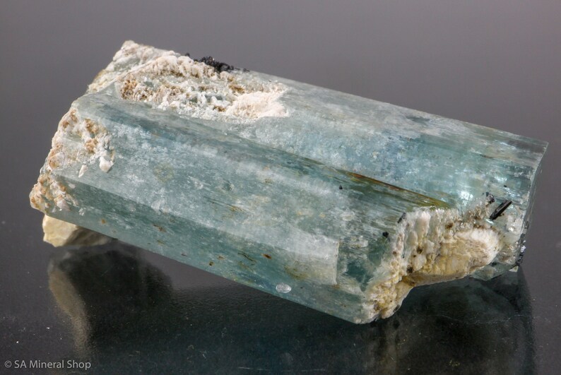 May include: A raw, aquamarine crystal with a rectangular shape. The crystal has a translucent, light blue hue with visible striations. White and beige mineral deposits are present on the edges and ends, with small black inclusions.