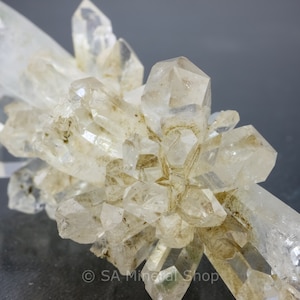 May include: A cluster of clear quartz crystals with a rough, natural texture. The crystals are arranged in a starburst pattern, with some of the points broken off. The crystals are set against a grey background.