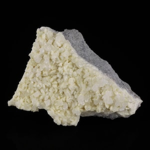 Może przedstawiać: Zbliżenie okazu mineralnego. Jedna strona ma jasnożółtą, krystaliczną strukturę, druga szarą, szorstką teksturę. Minerał jest umieszczony na czarnym tle.