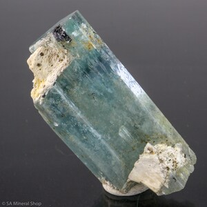 May include: A translucent, aquamarine-colored tourmaline crystal with a rectangular shape. The crystal has a slight blue-green hue and is partially encased in a white, textured matrix. Small black inclusions are visible.