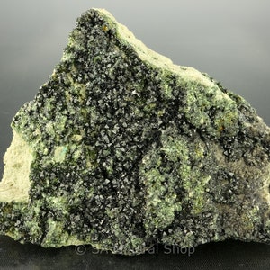 May include: A close-up of a rock specimen with black crystals embedded in a green matrix. The crystals are sharp and well-defined, and the matrix is a light green color. The rock is rough and textured.