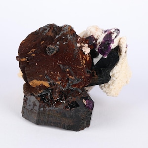 May include: A natural mineral specimen featuring a dark reddish-brown, rough-textured formation with black and purple crystalline structures. The specimen also includes areas of white and cream-colored mineral deposits.