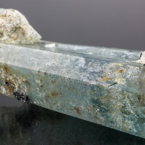 May include: A translucent, light blue aquamarine crystal with a rough, textured surface and a crystalline structure. The crystal has a rectangular shape with a lighter, rougher section at the top. Small black mineral inclusions are visible.
