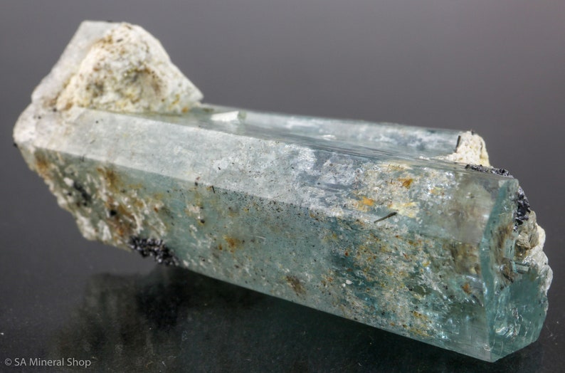 May include: A translucent, light blue aquamarine crystal with a rough, textured surface and a crystalline structure. The crystal has a rectangular shape with a lighter, rougher section at the top. Small black mineral inclusions are visible.
