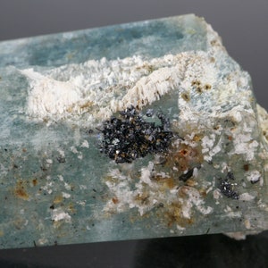 May include: A close-up of a raw aquamarine crystal with a pale blue hue. The crystal has a rough, textured surface with white and black mineral inclusions. The overall appearance is natural and unpolished.
