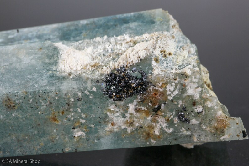 May include: A close-up of a raw aquamarine crystal with a pale blue hue. The crystal has a rough, textured surface with white and black mineral inclusions. The overall appearance is natural and unpolished.