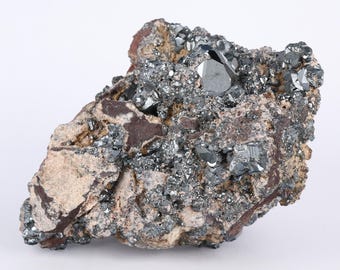 May include: A natural mineral specimen featuring a cluster of dark, metallic gray crystals embedded in a matrix of tan and reddish-brown rock. The crystals have sharp, geometric shapes, reflecting light. The overall texture is rough and uneven.