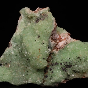 May include: A cluster of green, crystalline mineral specimens with a rough, textured surface. The mineral has a light green colour and is set against a black background.