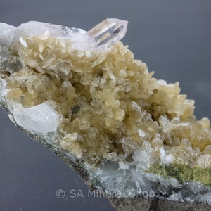 Op de afbeelding: Een cluster van lichtbruine kristallen met een helder kwartskristal bovenop. De kristallen bevinden zich op een donkergrijze rots met een groen mineraal.