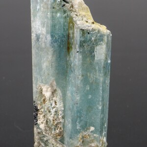 May include: A translucent, light blue aquamarine crystal with a columnar structure. The crystal has a rough, textured surface with areas of white and black mineral inclusions. The overall shape is a vertical, elongated prism.