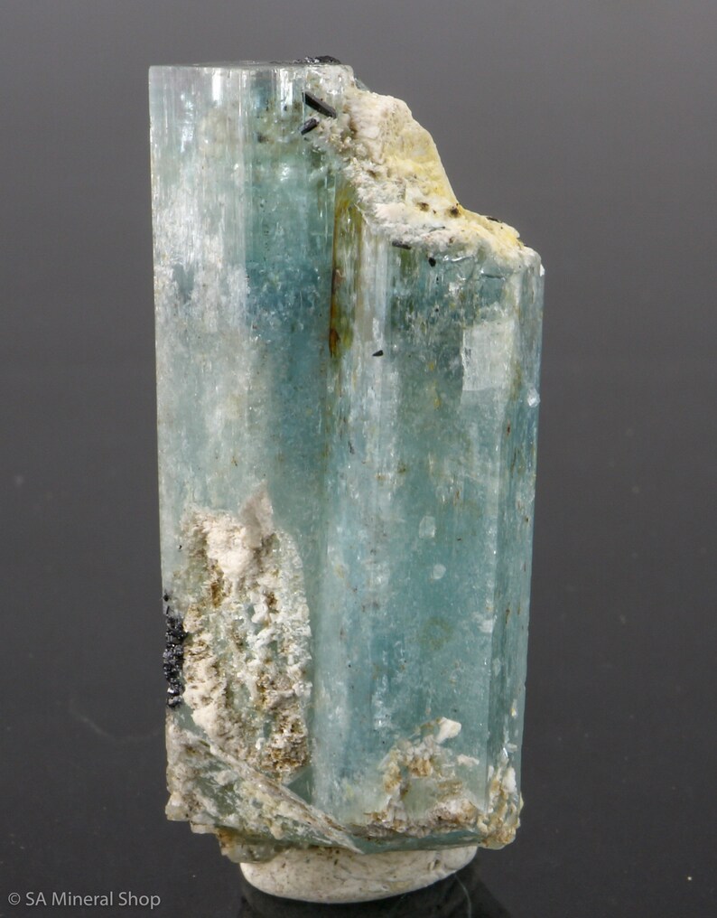 May include: A translucent, light blue aquamarine crystal with a columnar structure. The crystal has a rough, textured surface with areas of white and black mineral inclusions. The overall shape is a vertical, elongated prism.