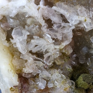 May include: Close-up of a mineral specimen featuring clusters of clear, crystalline structures. The crystals are set against a backdrop of off-white and pale yellow hues, with hints of green and brown.