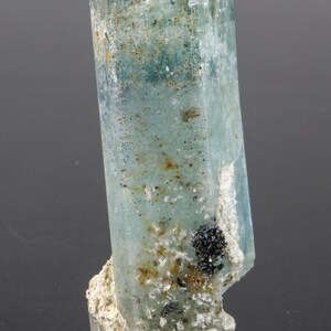 May include: A translucent, light blue aquamarine crystal with a rectangular shape. The crystal has a slightly rough texture and is mounted on a white, textured base. Small black mineral deposits are visible on the crystal and base.