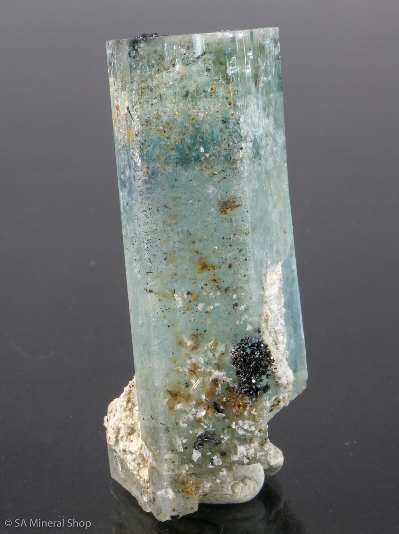 May include: A translucent, light blue aquamarine crystal with a rectangular shape. The crystal has a slightly rough texture and is mounted on a white, textured base. Small black mineral deposits are visible on the crystal and base.