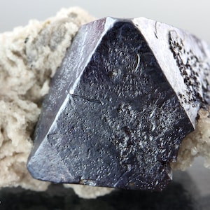 May include: A large, dark gray, metallic, octahedral crystal of pyrite, with a slightly iridescent sheen, is partially embedded in a matrix of white, fibrous calcite crystals.