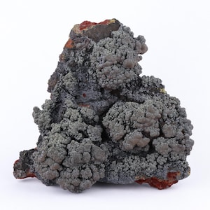May include: A close-up of a mineral specimen with a textured, gray surface. The mineral has a clustered, bubbly appearance with reddish-brown accents at the base and top. The background is plain white.