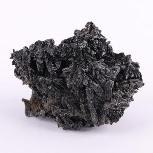 May include: A close-up of a dark, textured mineral specimen. The rock is primarily black with a rough, crystalline surface. The mineral appears to be a cluster of needle-like formations, creating a spiky appearance. The background is plain white.