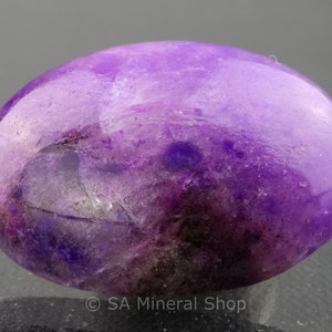May include: A smooth, oval-shaped purple amethyst gemstone with a light purple hue and darker purple inclusions. The gemstone is polished and has a glossy finish.