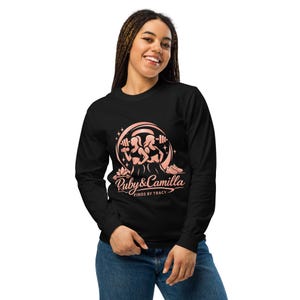 Puede incluir: Una camiseta negra de manga larga con un estampado gráfico en rosa claro de dos figuras levantando pesas, una barra de pesas y zapatillas deportivas. El texto Ruby & Camilla FINDS BY TRACY aparece en el pecho con una fuente estilizada.