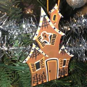 May include: A brown gingerbread house-shaped Christmas ornament with white trim and black details. The ornament features a ghost, spiderwebs, and a fence.