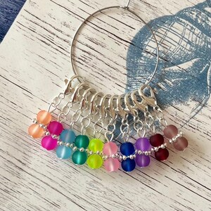 May include: A set of 10 colorful stitch markers with silver clasps attached to a silver ring. The markers are made of frosted glass beads and are arranged in a rainbow pattern.