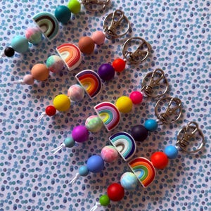May include: A collection of rainbow-themed beaded keyrings, featuring a variety of colourful beads and rainbow-shaped charms. Each keyring has a silver-coloured clasp. The keyrings are arranged on a blue and white polka dot background.