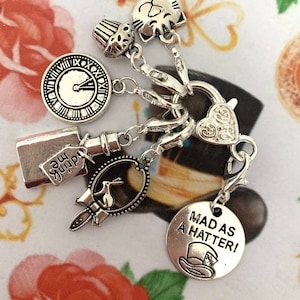 May include: Silver charm bracelet featuring Alice in Wonderland themed charms, including a clock, a cupcake, a cat, a bottle, a mirror, and a charm that says "Mad as a Hatter!"