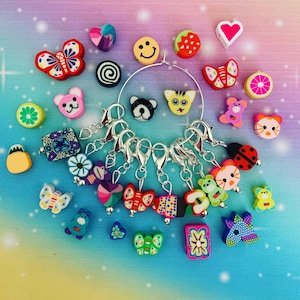 May include: A set of 10 colourful, handmade polymer clay stitch markers with various designs, including butterflies, animals, and geometric shapes. The markers are attached to silver-toned metal rings.