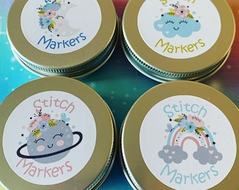 May include: Four gold containers with white lids, each featuring a different colourful design with the text "Stitch Markers". The designs include a crescent moon, a cloud, a planet, and a rainbow.