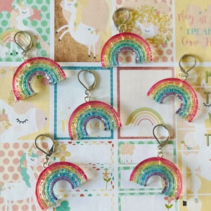 May include: Five pairs of rainbow-coloured earrings with silver hooks. Each earring is shaped like a rainbow with glitter embedded in the resin.
