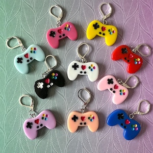 May include: A selection of colourful video game controller keyrings. Each keyring features a miniature controller in various colours, including pink, yellow, red, blue, and black, with silver-tone metal hardware. The controllers have heart and button details.