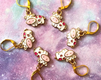 May include: Six gold-tone cow charm earrings with pink and white enamel. Each charm features a white cow with pink spots and a pink nose.