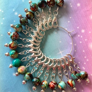 May include: A set of 15 silver stitch markers with turquoise and brown marbled glass beads. The markers are attached to a silver ring.