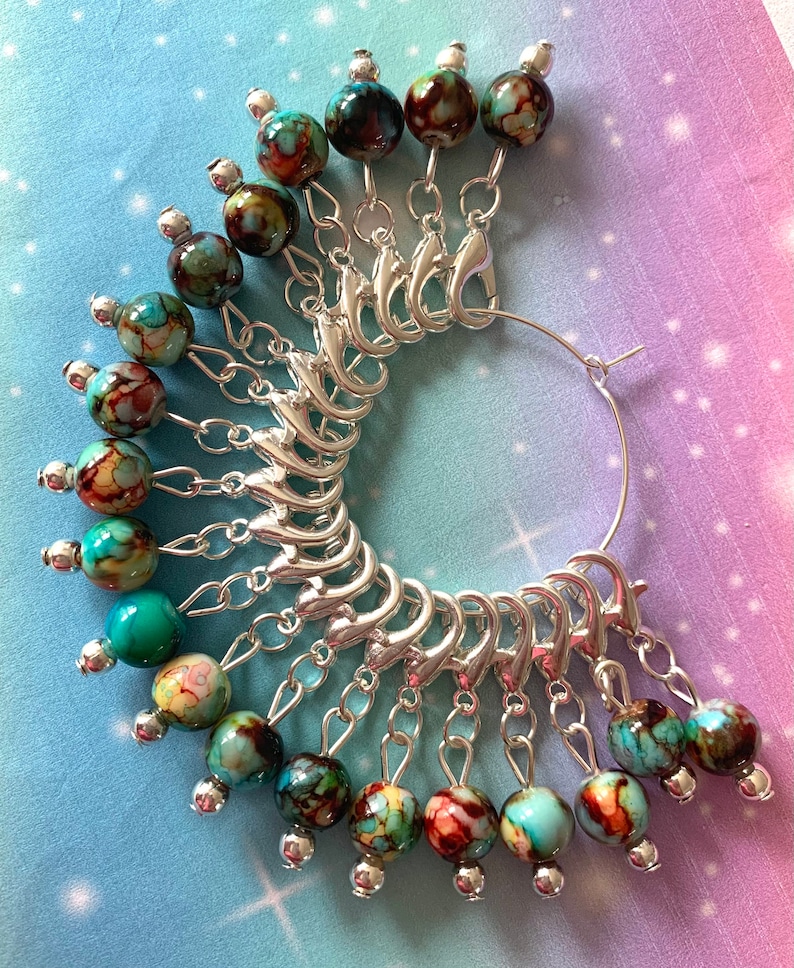 May include: A set of 15 silver stitch markers with turquoise and brown marbled glass beads. The markers are attached to a silver ring.