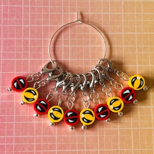 May include: A set of 10 stitch markers with a silver hoop. The markers are red and yellow with a black design.