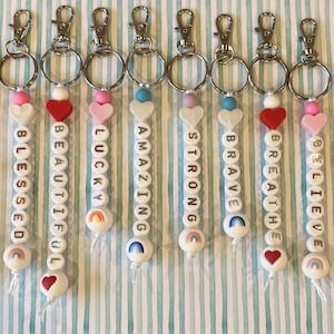 May include: Ten colourful keychains with positive messages, each featuring a heart and a rainbow charm. The messages include "Got This", "Blessed", "Beautiful", "Lucky", "Amazing", "Strong", "Brave", "Breathe", "Believe", and "I'm Enough".