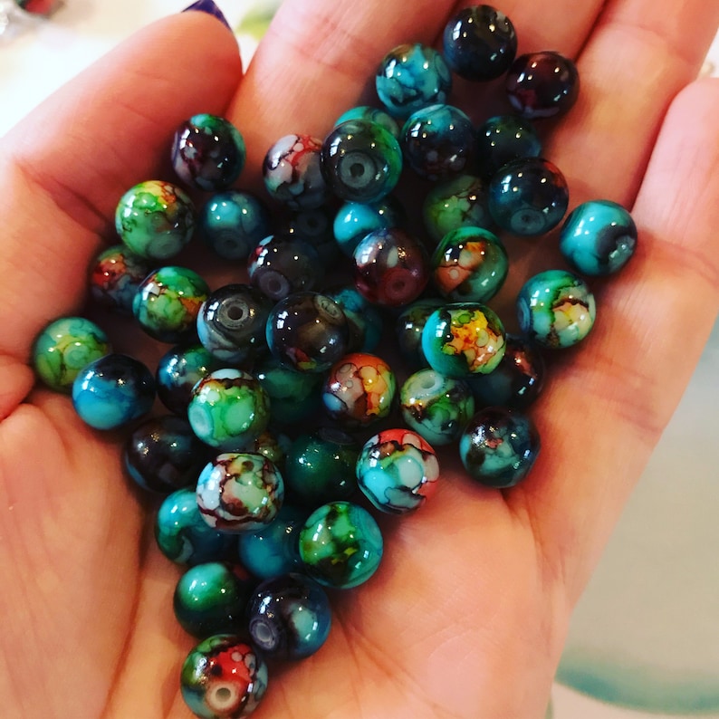 May include: A collection of colourful glass beads with swirling patterns in shades of blue, green, and black. The beads are round and have a glossy finish.