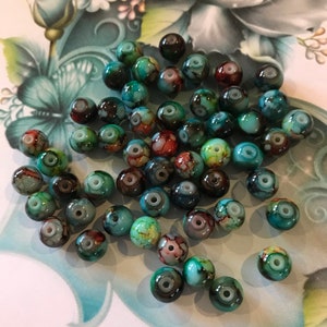 May include: A collection of round, turquoise, green, and brown glass beads with white swirls. The beads have a marbled effect and are scattered on a white surface with a floral design.