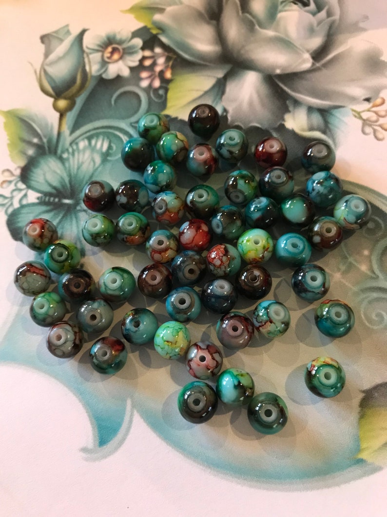 May include: A collection of round, turquoise, green, and brown glass beads with white swirls. The beads have a marbled effect and are scattered on a white surface with a floral design.